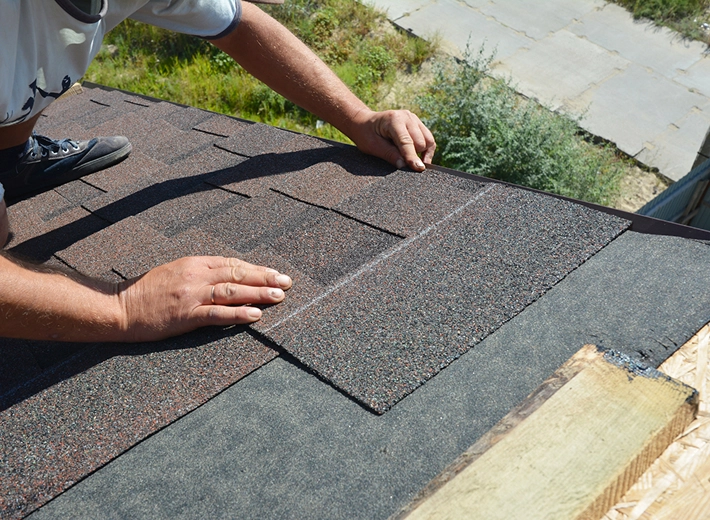 Person installing asphalt shingles on a sloped roof, using their hands to align the shingles over roofing felt.